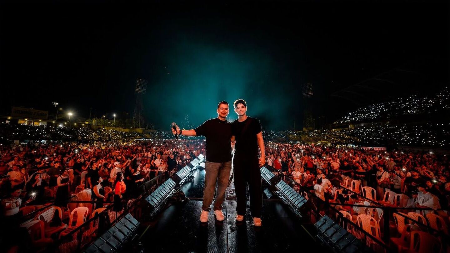 Dos artistas de frente en el escenario, con el estadio lleno detrás