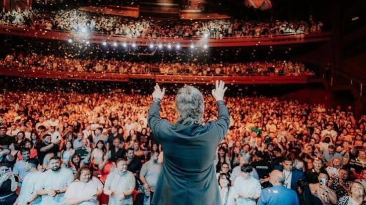 Un hombre de espaldas en el escenario, con los brazos en alto, frente a un teatro lleno