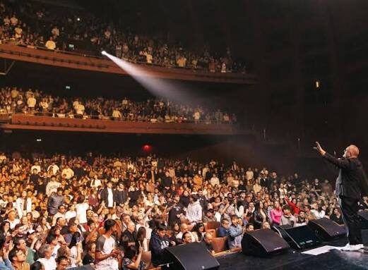 Teatro lleno con un reflector encendido y, en el escenario, una persona con el brazo extendido